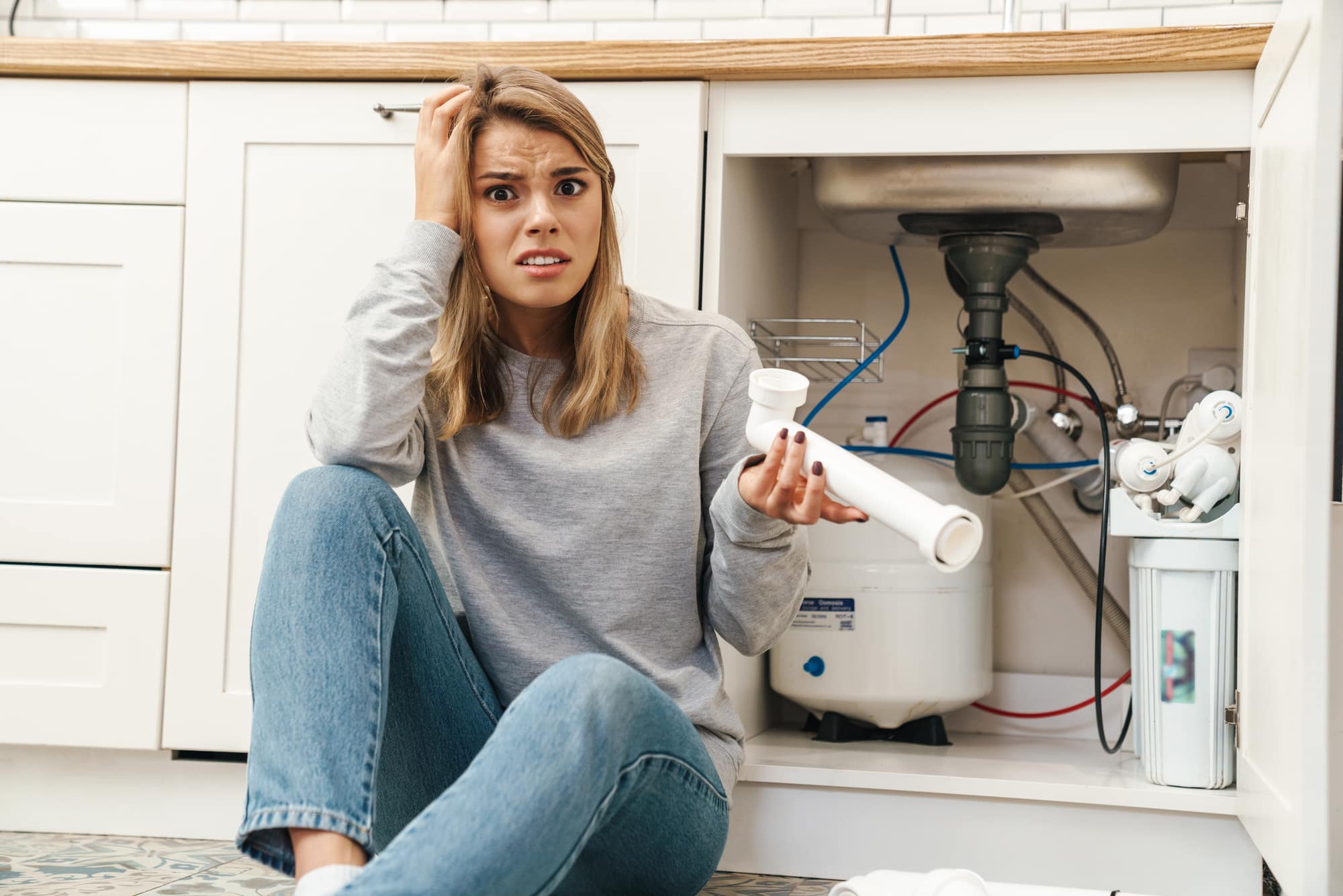 Photo of displeased confused blonde woman with plumbing pipes while sitting on floor near kitchen sink